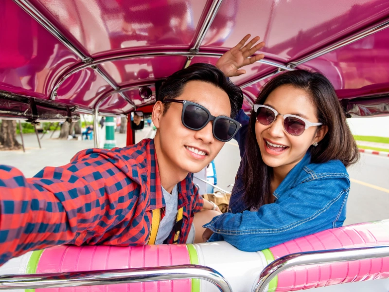 Two people sitting inside a pink tuk-tuk with green and white accents, one taking a selfie; faces are blurred for privacy.