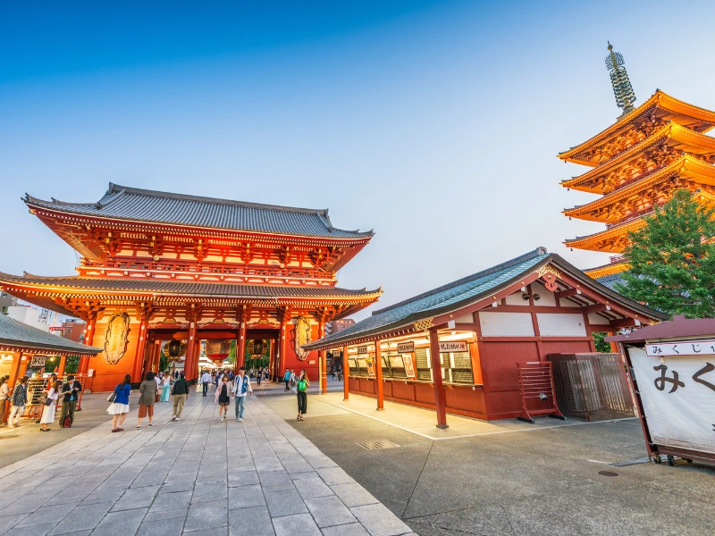 Senso-Ji Temple in Tokyo illuminated with warm lights, featuring traditional architecture, a five-story pagoda, and visitors walking toward the main gate under a clear blue sky.