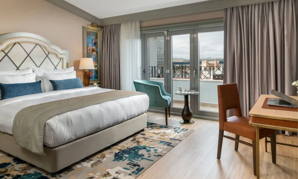 Elegant hotel room at Millennium Istanbul Golden Horn featuring a king-size bed with white linens and blue accent pillows, modern decor, a work desk, and a balcony with city views.