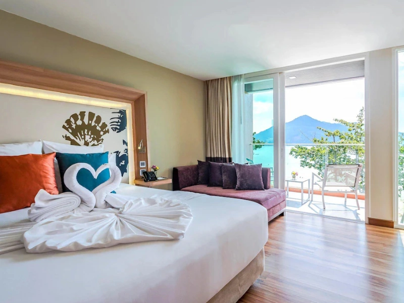 A well-lit, modern bedroom with a large bed that has white linens and decorative pillows. Two towel swans forming a heart shape are placed on the bed. The room has wooden flooring and features a large window or sliding door that opens to a balcony with outdoor furniture. The view from the balcony includes greenery and mountains in the distance.