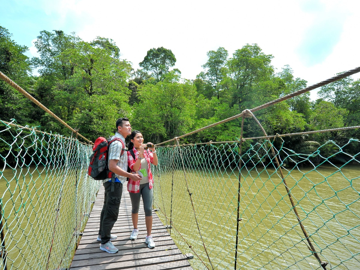 Two people standing on a wooden suspension bridge with netted sides, suspended over water and surrounded by lush green trees, wearing casual clothing and backpacks.