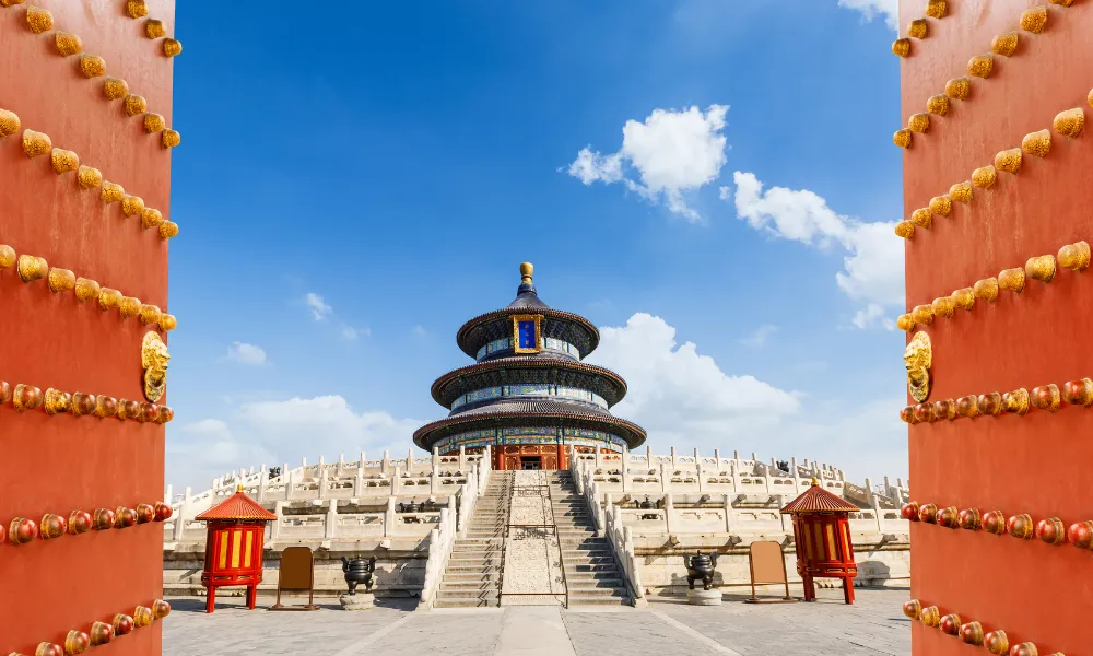 The Temple of Heaven in Beijing, China, viewed through an open red gate adorned with golden knobs. The temple features a circular, three-tiered structure with a blue roof and intricate details. The sky is clear with a few clouds.