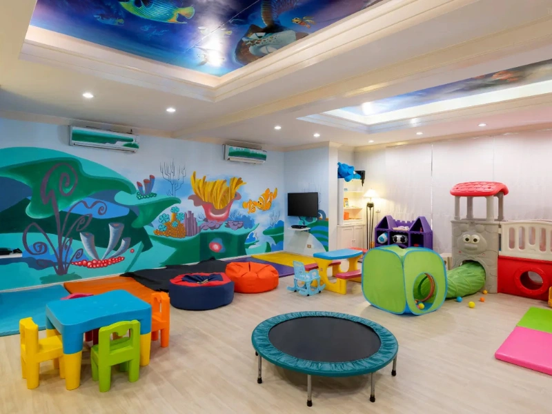 Colorful children's playroom with underwater-themed wall and ceiling art, featuring a trampoline, playhouse with slides, tunnel, bean bags, and small tables with chairs.