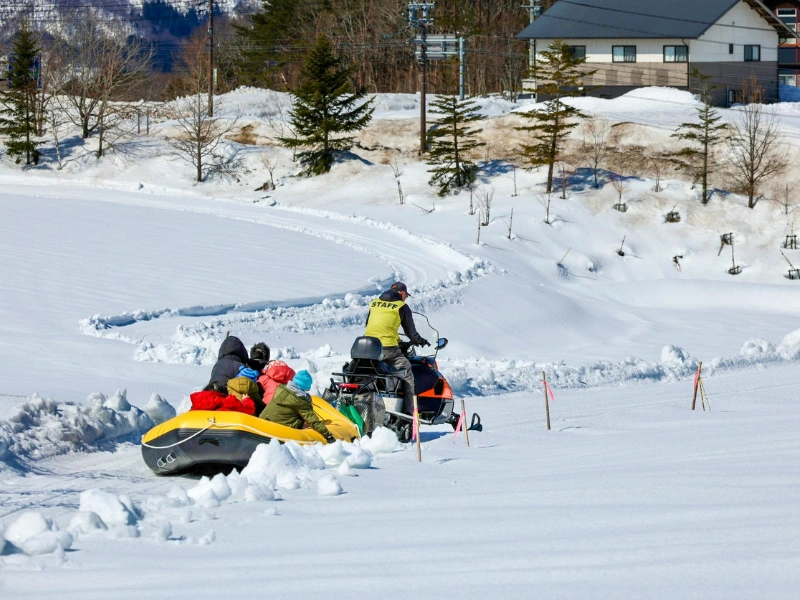 Group of people seated in an inflatable raft being pulled by a snowmobile along a snowy path in Hokkaido, with snow-covered ground, trees, and a building in the background