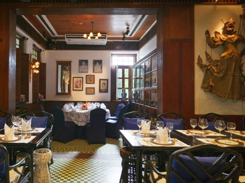 The image shows the interior of a restaurant with a cozy and elegant ambiance. The room features wooden paneling, decorative wall art, and framed pictures. There are several tables set with white tablecloths, blue chairs, and neatly arranged tableware including glasses, plates, and napkins. The floor has a checkered pattern in shades of green and yellow. A large window allows natural light to enter the room.