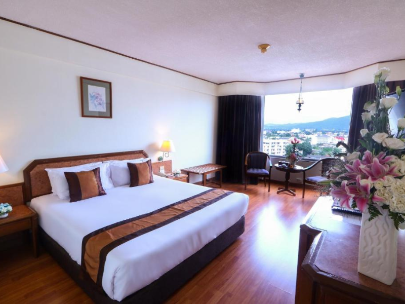 Well-furnished hotel room with a large bed, bedside tables and lamps, framed artwork above the bed, a window with city and mountain views, a small table with two chairs, a dresser with flowers, and wooden flooring.