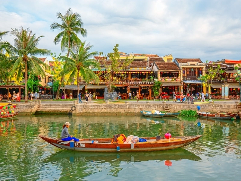 Scenic riverside view with boats, palm trees, and traditional buildings adorned with colorful decorations and lanterns, featuring people walking along the river and a person in a boat under a partly cloudy sky.