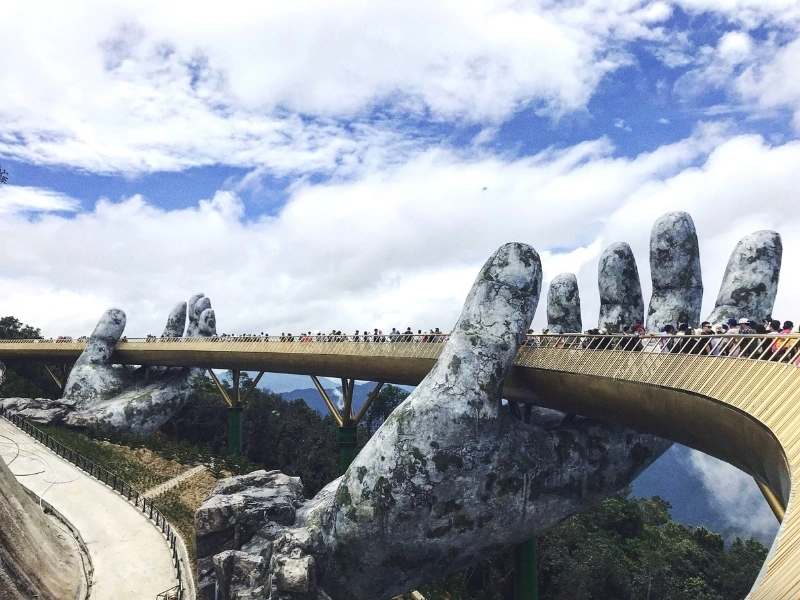 Golden Bridge in Da Nang, Vietnam, supported by two giant stone hands, spanning a lush green landscape with mountains in the background and people walking along the bridge under a partly cloudy sky.