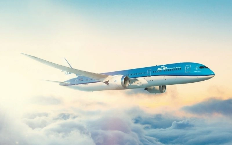 A KLM Royal Dutch Airlines Boeing 787 Dreamliner flying above the clouds during sunrise, with a clear blue sky in the background.