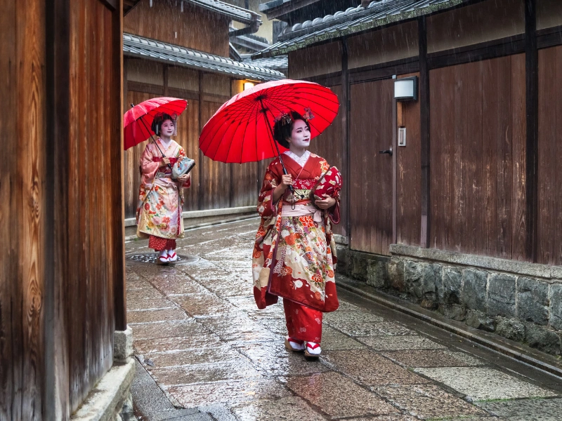 Two individuals in traditional kimonos holding red umbrellas walking down a narrow, wet street in Kyoto, lined with wooden buildings