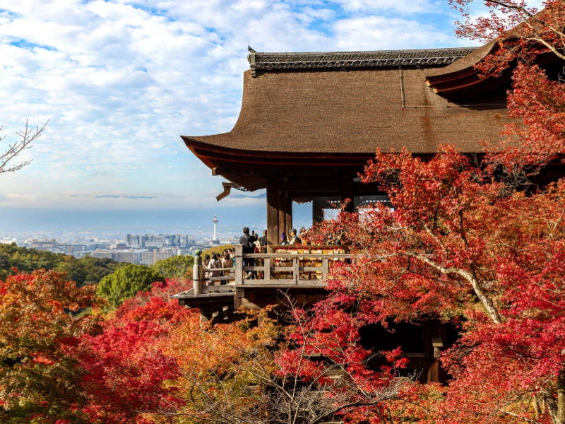 Kiyomizu-dera Temple in Kyoto, Japan, perched on a hillside with a large wooden stage overlooking vibrant autumn foliage and a distant cityscape under a partly cloudy sky.