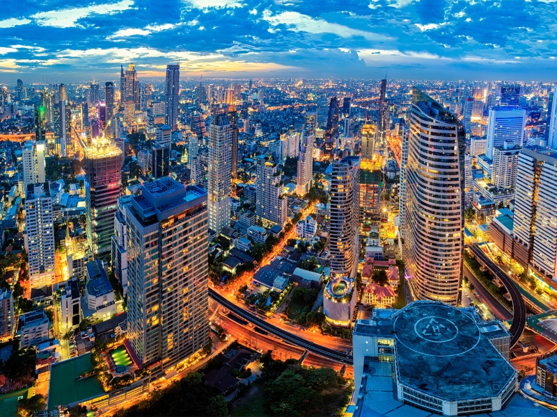 Vibrant cityscape at dusk or dawn featuring illuminated high-rise buildings, a partly cloudy sky with blue and orange hues, and a prominent skyscraper with a circular rooftop structure in the foreground.