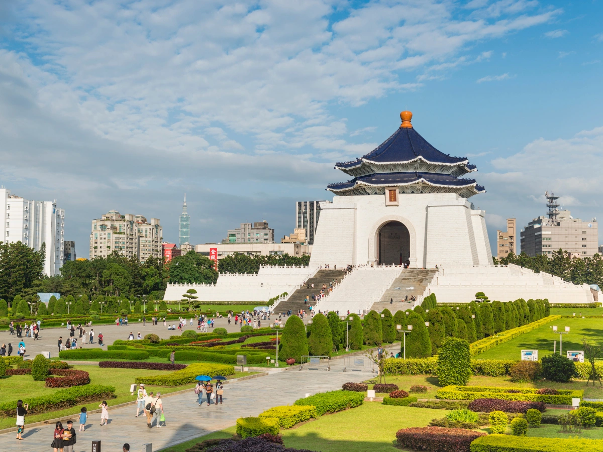 The Chiang Kai-shek Memorial Hall in Taipei, Taiwan, featuring traditional Chinese architecture with a blue-tiled roof and white walls, surrounded by gardens, pathways, and visitors, with modern buildings and a partly cloudy blue sky in the background.