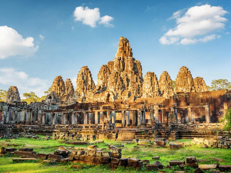 The ancient Bayon temple complex in Angkor Thom, Cambodia, featuring stone towers with carved faces, intricate architectural details, scattered ruins, and a grassy foreground under a partly cloudy blue sky.