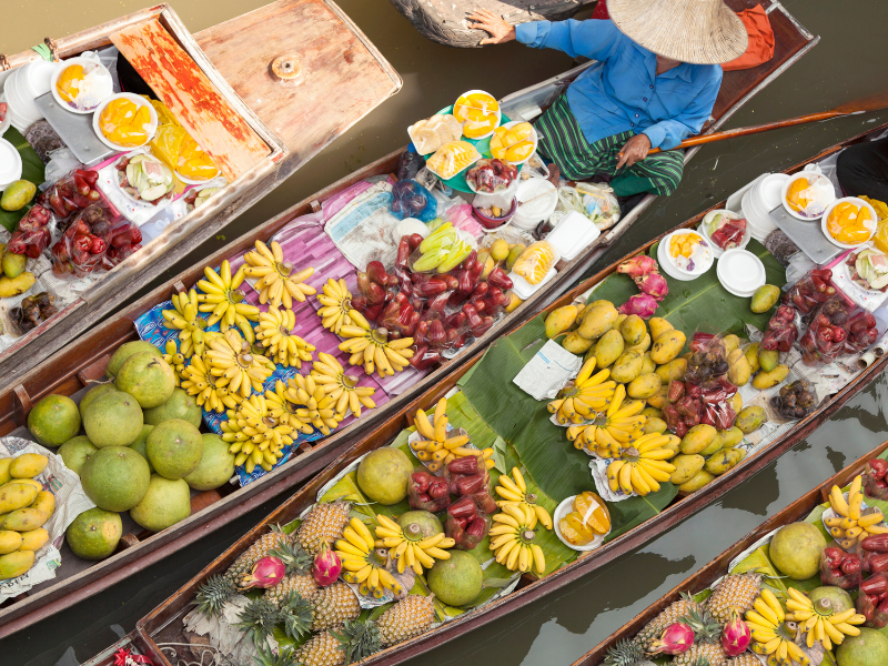 The image shows a vibrant floating market with several boats filled with various fruits and goods. The boats are packed closely together on the water, and each boat is loaded with an assortment of colorful fruits such as bananas, pineapples, mangoes, and other tropical produce. There are also containers of what appears to be food or drinks. One person wearing a conical hat and blue shirt is visible in one of the boats.