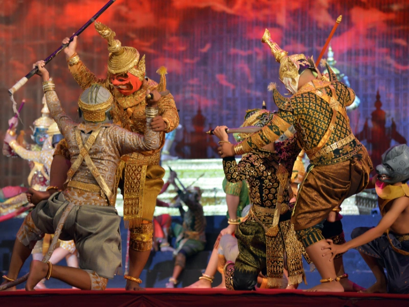 A group of performers in traditional Thai costumes act out a dramatic scene with swords on a vibrant stage filled with red and purple lighting.