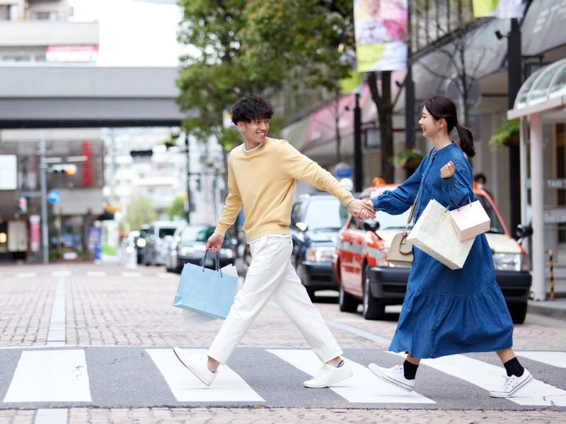 Couple walking hand-in-hand across a city crosswalk, dressed casually and carrying shopping bags, with blurred faces for privacy. Urban background includes parked cars, storefronts, trees, and colorful banners.