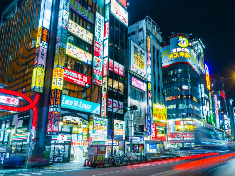 Bustling Tokyo street at night, lit up by vibrant neon signs and colorful billboards in Japanese, with busy traffic and pedestrians creating a lively urban atmosphere.