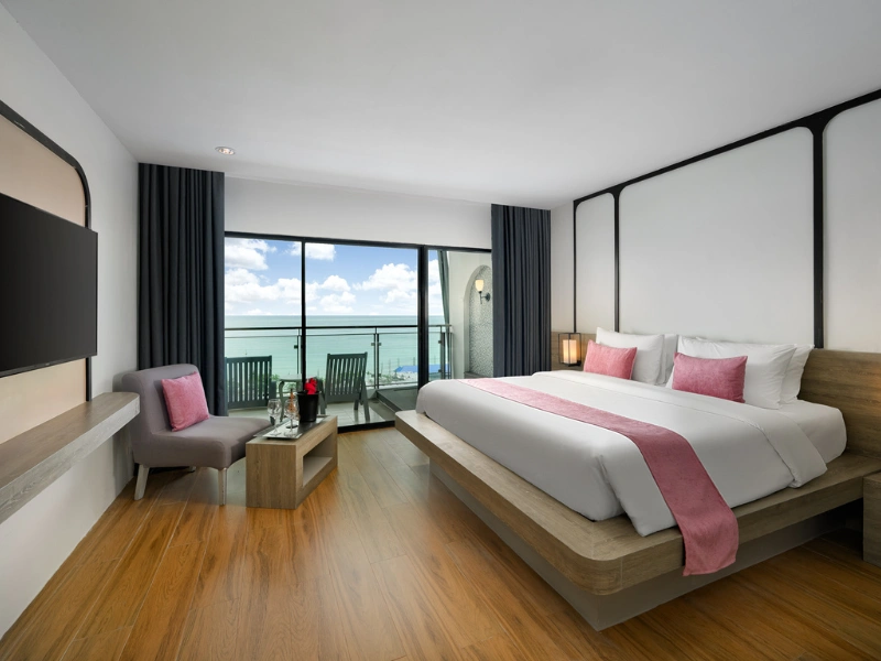 A modern, well-lit hotel room with a large bed that has white linens and pink accents, including pillows and a runner. There is a small seating area with a grey chair and pink cushion, along with a small table holding flowers and what appears to be wine glasses. The room features wooden flooring, dark curtains framing large windows or sliding doors that open to a balcony with chairs, offering an ocean view.