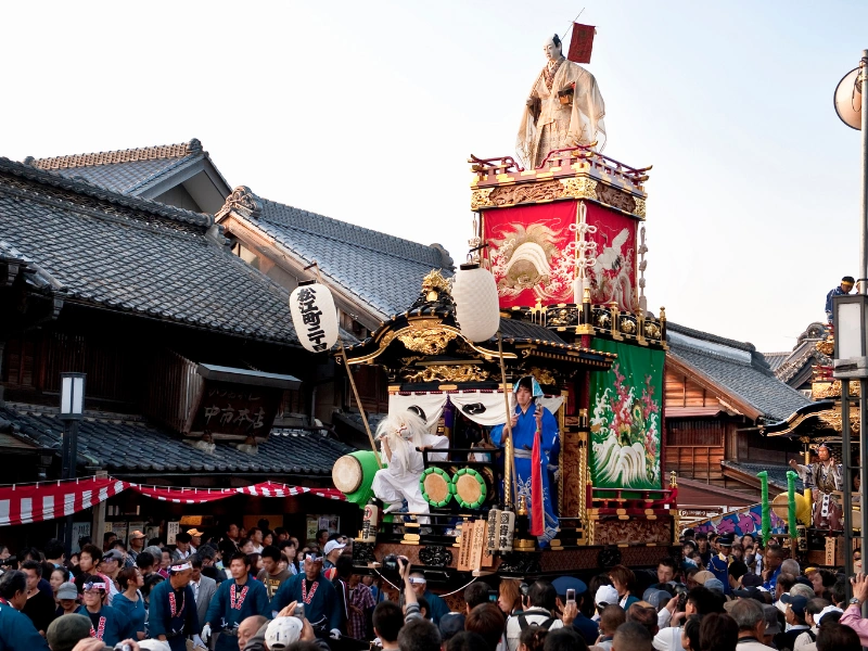 Traditional Japanese float decorated with lanterns, banners, and figures being paraded through a street during the Kawagoe Festival, with crowds of people and traditional buildings in the background.