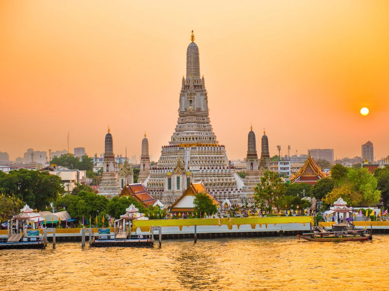 Wat Arun temple in Bangkok with its ornate central prang silhouetted against an orange sunset sky, viewed from across the Chao Phraya River.