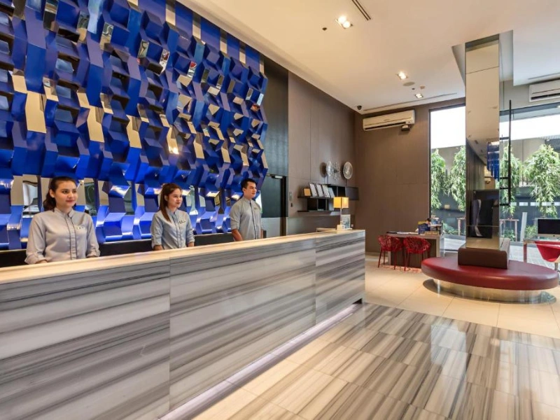 Modern hotel reception area with three receptionists behind a long marble-patterned counter, set against a stylish wall with blue and metallic accents—welcoming for families and children.