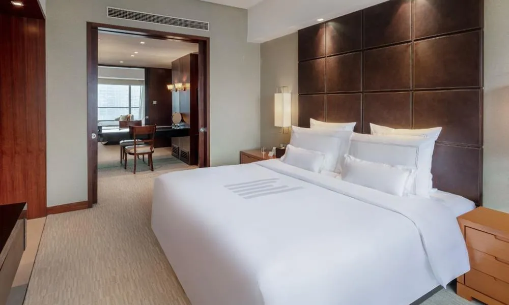 Elegant suite at Grand Millennium Beijing featuring a king-size bed with crisp white linens, leather-paneled headboard, bedside tables, and a separate living area with city views.