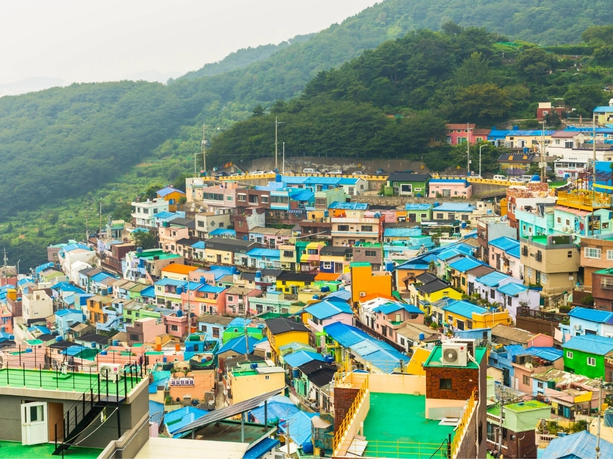 Densely packed hillside village with colorful rooftops in shades of blue, green, yellow, and orange, surrounded by lush green hills and vegetation. The image highlights urban density and architectural diversity in a mountainous landscape.