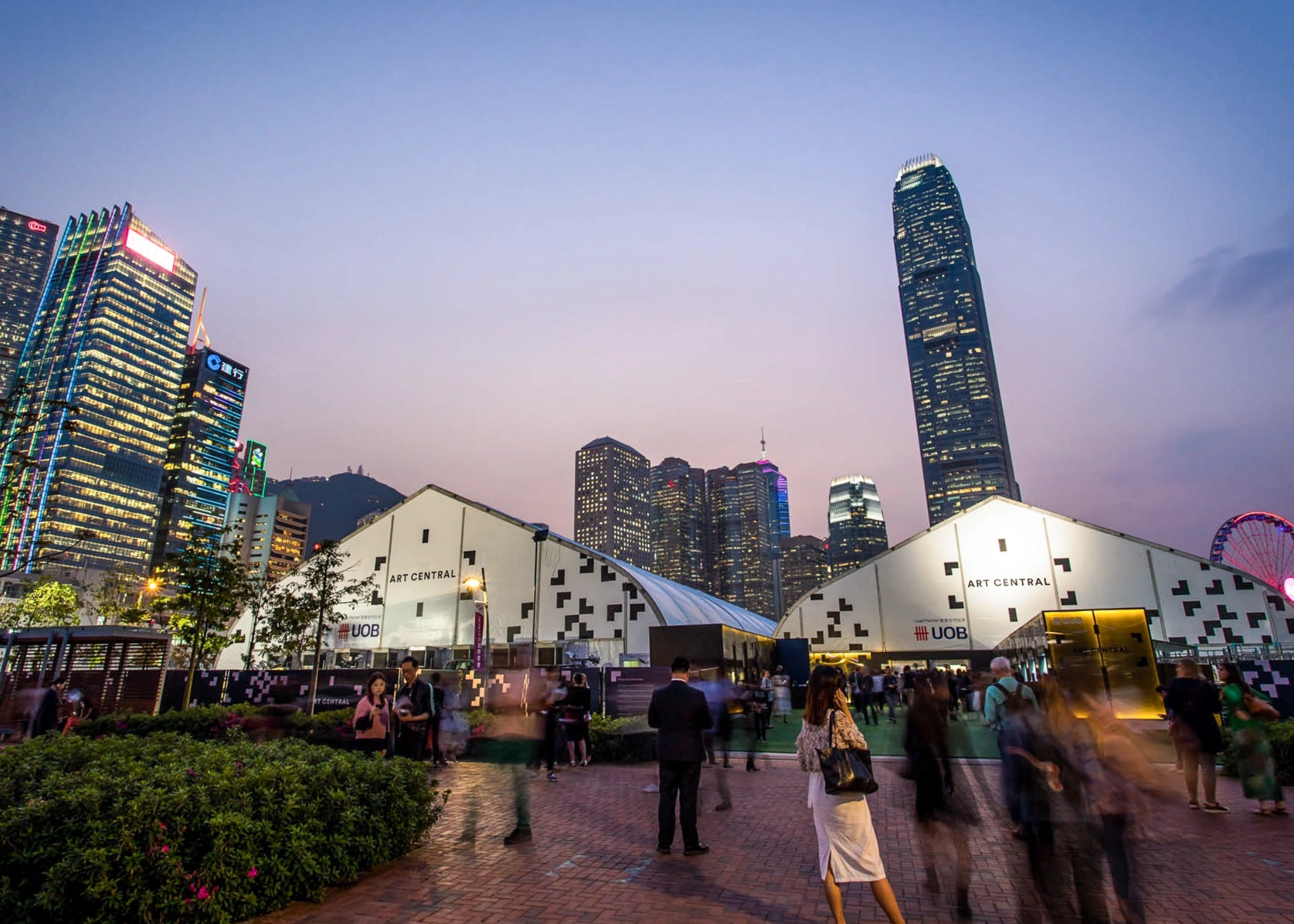 Twilight outdoor scene with people walking near two large white tents labeled 'ART CENTRAL' and 'UBS', set against a backdrop of illuminated skyscrapers and a gradient sky transitioning from light blue to pink.
