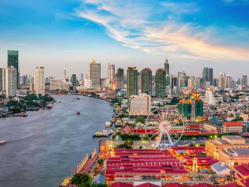 Vibrant cityscape with a river, high-rise buildings, a large Ferris wheel, and colorful structures under a partly cloudy sky at sunrise or sunset.