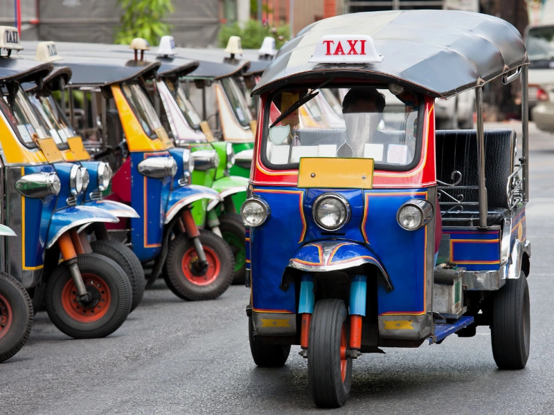 Group of colorful tuk-tuks parked together, with a blue tuk-tuk in the foreground featuring red and yellow accents and a 'TAXI' sign on top.