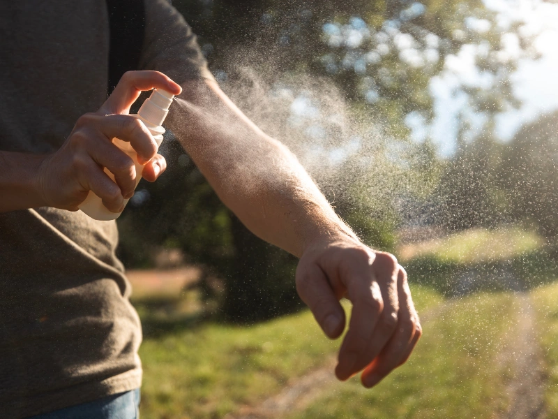 Person spraying an aerosol can on their forearm in a sunlit outdoor setting with greenery, possibly applying insect repellent in Thailand.